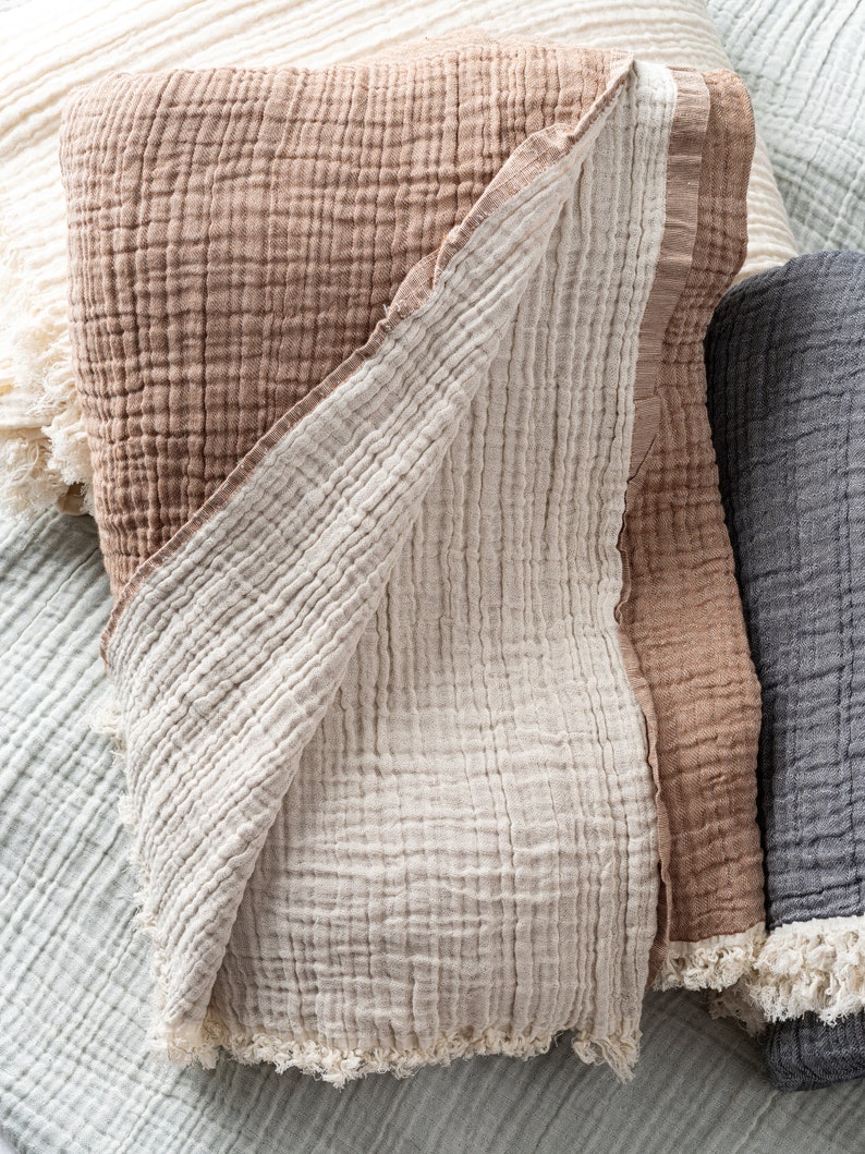 May include: A folded muslin blanket featuring a textured, crinkled fabric in shades of tan and cream. The blanket has a visible grid-like weave pattern and frayed, raw edges along the bottom hem, layered among other similar blankets in various colours.