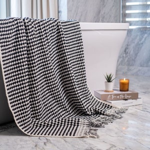 May include: A black and white woven throw blanket featuring a repeating geometric dot pattern and fringed edges. The blanket is draped over the side of a white bathtub, showcasing a textured knit construction with a solid cream-colored border.