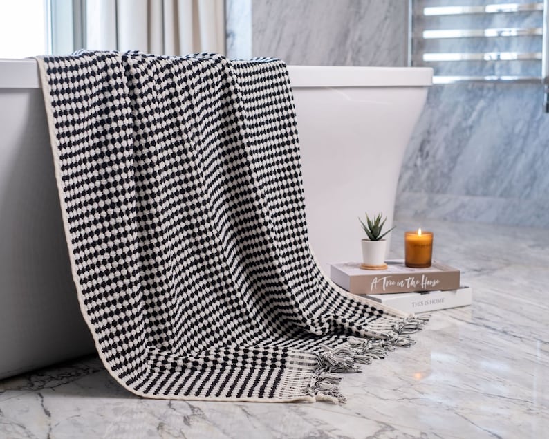 May include: A black and white woven throw blanket featuring a repeating geometric dot pattern and fringed edges. The blanket is draped over the side of a white bathtub, showcasing a textured knit construction with a solid cream-colored border.