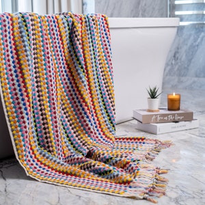 May include: A rectangular bath towel featuring a vibrant, multi-colored textured pattern of small raised dots in shades of red, blue, yellow, pink, and grey. The towel has a cream-colored border and decorative fringe along the bottom edge.