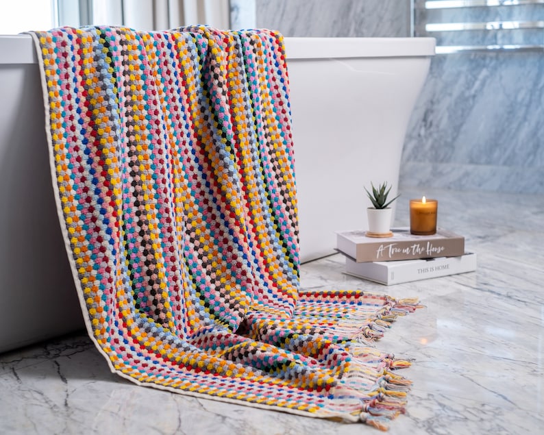 May include: A rectangular bath towel featuring a vibrant, multi-colored textured pattern of small raised dots in shades of red, blue, yellow, pink, and grey. The towel has a cream-colored border and decorative fringe along the bottom edge.