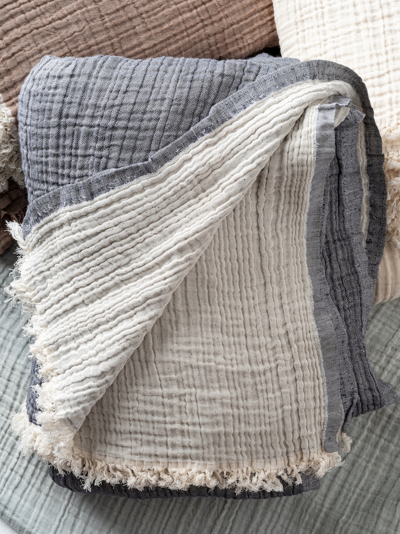 May include: A folded multi-layered muslin blanket featuring a dark charcoal grey exterior and a cream-coloured interior. The fabric has a distinct crinkled, textured grid pattern and is finished with frayed, raw edges along the border.