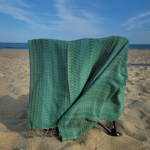 May include: A green woven beach chair with a metal frame, set up on a sandy beach with a blue sky and ocean in the background.