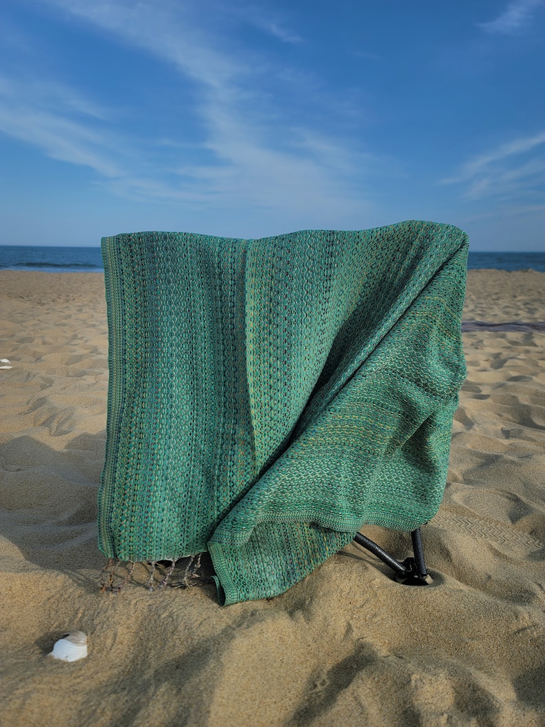 May include: A green woven beach chair with a metal frame, set up on a sandy beach with a blue sky and ocean in the background.