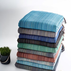 May include: A stack of ten woven cotton throws in various shades of blue, green, and brown. The throws are folded and stacked on a white surface.
