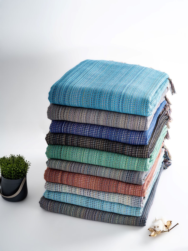 May include: A stack of ten woven cotton throws in various shades of blue, green, and brown. The throws are folded and stacked on a white surface.