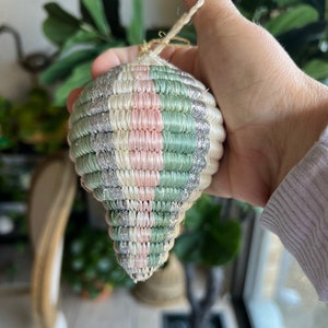 May include: A woven Christmas ornament with a pink, green, and silver striped pattern. The ornament is made of natural fibers and has a rustic, handmade look.