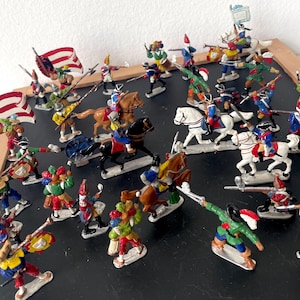 Puede incluir: Una colección de soldaditos de juguete antiguos que representan una escena de batalla. Las figuras están pintadas en varios colores, incluyendo rojo, azul, verde y amarillo. Algunos soldados están montados a caballo y se ven banderas. Los juguetes están colocados sobre una superficie negra.