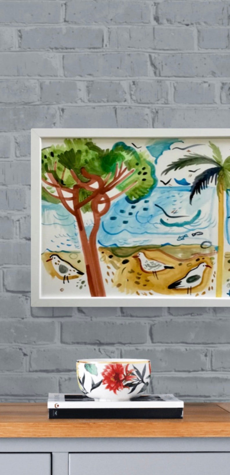 May include: A framed watercolor painting of a beach scene with two palm trees, blue water, and white birds. The painting is in a white frame and is hanging on a white brick wall.