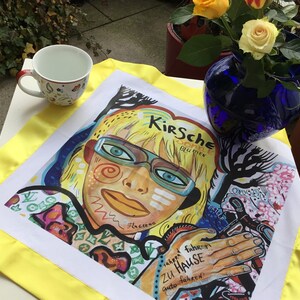 Pode incluir: Uma toalha de cozinha branca com uma borda amarela apresenta uma ilustra&ccedil;&atilde;o colorida de uma pessoa com um rosto amarelo e vermelho brilhante, usando &oacute;culos e uma camisa vermelha e verde. O texto "Kirsche Blume" &eacute; vis&iacute;vel no canto superior direito da imagem. A toalha de cozinha &eacute; uma &oacute;tima adi&ccedil;&atilde;o a qualquer cozinha.
