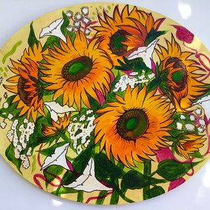 Könnte beinhalten: Eine handbemalte ovale Platte mit gelbem Hintergrund zeigt einen Strauß Sonnenblumen, weiße Mondblumen und kleine weiße Blüten. Die Sonnenblumen haben gelbe Blütenblätter und braune Zentren mit grünen Blättern. Die Mondblumen sind weiß mit einem gelben Zentrum. Die kleinen weißen Blüten sind zu einem Büschel zusammengefasst.