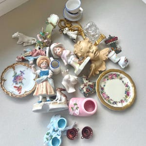 Könnte beinhalten: Eine Sammlung von dekorativen Miniaturgegenständen, darunter Porzellanteller mit Blumenmustern, Keramikfiguren von Kindern und Tieren, ein goldfarbener Cherubkopf, ein rosa Babyschuh, blaue Babyschuhe mit Schleifen und kleine Metallformen.