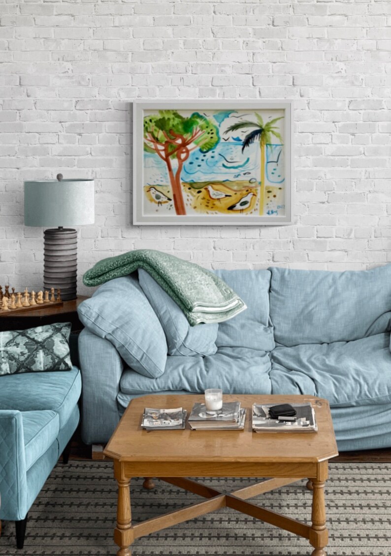 May include: A light blue sofa with a green throw blanket, a white lamp with a gray base, and a wooden coffee table with books and a glass on top. A framed watercolor painting of a beach scene with palm trees and birds hangs on a white brick wall.