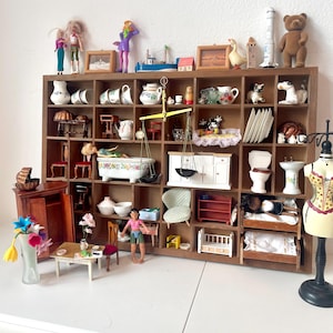 May include: A wooden display cabinet filled with miniature dollhouse items. The cabinet contains dolls, crockery, a bathtub, scales, a bed, and a mannequin. The cabinet is brown and sits on a white surface.