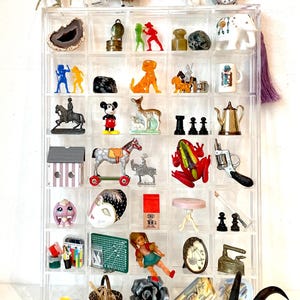 May include: A clear acrylic display case filled with a variety of miniature figurines, toys, and trinkets. The case has 36 compartments and is filled with items such as a rocking horse, a deer, a frog, a doll, a teapot, a clock, a house, a mask, a basket, a rose, and a skull.