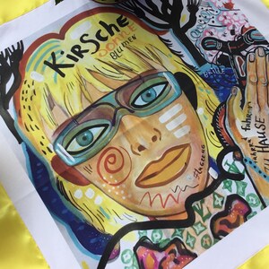 Pode incluir: Um quadrado de tecido branco com uma borda amarela. O tecido apresenta uma ilustra&ccedil;&atilde;o colorida de uma mulher com cabelo loiro usando &oacute;culos. O texto "Kirsche" est&aacute; escrito em preto acima da cabe&ccedil;a da mulher. O texto "donne" est&aacute; escrito em preto abaixo da cabe&ccedil;a da mulher. O texto "blu mew" est&aacute; escrito em preto abaixo do texto "donne".