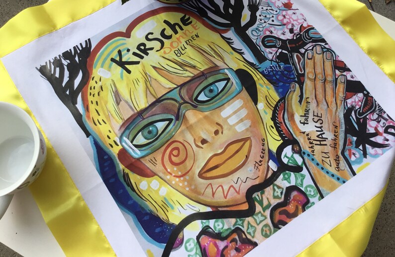 Pode incluir: Um quadrado de tecido branco com uma borda amarela. O tecido apresenta uma ilustra&ccedil;&atilde;o colorida de uma mulher com cabelo loiro usando &oacute;culos. O texto "Kirsche" est&aacute; escrito em preto acima da cabe&ccedil;a da mulher. O texto "donne" est&aacute; escrito em preto abaixo da cabe&ccedil;a da mulher. O texto "blu mew" est&aacute; escrito em preto abaixo do texto "donne".