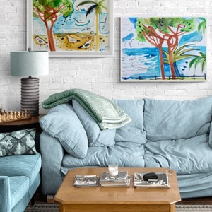 May include: Two framed watercolor paintings of seascapes with palm trees and blue water. The paintings are hanging on a white brick wall above a light blue sofa with a green throw blanket. A wooden coffee table with magazines and a candle sits in front of the sofa.