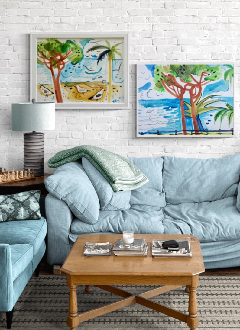 May include: Two framed watercolor paintings of seascapes with palm trees and blue water. The paintings are hanging on a white brick wall above a light blue sofa with a green throw blanket. A wooden coffee table with magazines and a candle sits in front of the sofa.