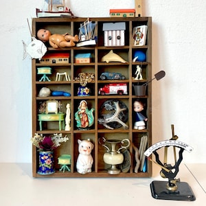 May include: A wooden drawer organizer with various small items, including a doll, a toy car, a small house, a ship, a vase, a pig figurine, a small mirror, a seashell, a piece of driftwood, a small scale, and a small bucket. The drawer organizer is made of dark wood and has 18 compartments.