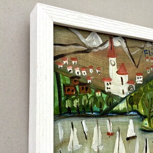 May include: A framed painting of a village scene with a church, houses, and trees. The painting is in a white frame and has a signature in the upper right corner that reads "Flueli".