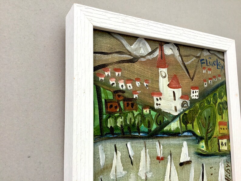 May include: A framed painting of a village scene with a church, houses, and trees. The painting is in a white frame and has a signature in the upper right corner that reads "Flueli".