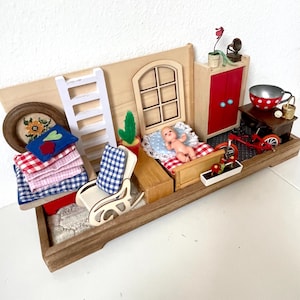 Pode incluir: Um conjunto de quarto de casa de bonecas em miniatura com uma moldura de madeira, um armário vermelho, uma cadeira de balanço, uma escada, uma cama com uma boneca, um cacto em vaso, uma janela, uma carroça vermelha, uma tigela e uma mesa redonda com uma xícara de chá e um bule.
