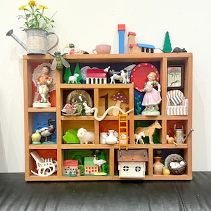 Könnte beinhalten: Eine hölzerne Schattenbox mit mehreren Fächern, gefüllt mit Miniaturfiguren, Tieren und Haushaltsgegenständen. Die Box ist in verschiedenen Farben bemalt, darunter Rot, Blau, Grün und Gelb. Die Box ist gefüllt mit einer Vielzahl von Gegenständen, darunter ein Miniaturhaus, ein Schaukelstuhl, eine Gießkanne, eine Gans, ein Schaf, ein Pferd, eine Ente, ein Teller, eine Tasse, ein Pilz, eine Leiter, eine Gießkanne, ein Vogel, ein Baum, ein Teller, eine Puppe, eine Fliege und eine Maus.