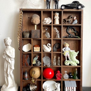 Puede incluir: Una estantería de madera con varias figuras y objetos en miniatura. Incluye una estatua, una libélula, un coche, figuras de hombres, una escultura de cabeza, una rosa, un cisne, un pájaro, una bola roja y otros objetos decorativos.