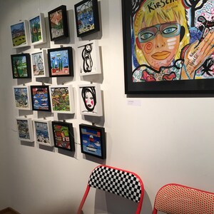 Pode incluir: Uma parede de galeria com obras de arte emolduradas que apresentam pinturas e ilustra&ccedil;&otilde;es coloridas. As obras incluem um retrato de uma mulher com &oacute;culos azuis e um fundo colorido, e v&aacute;rias pe&ccedil;as emolduradas menores com paisagens e paisagens marinhas.