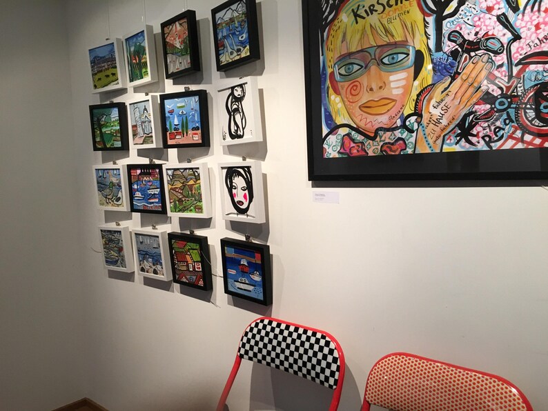 Pode incluir: Uma parede de galeria com obras de arte emolduradas que apresentam pinturas e ilustra&ccedil;&otilde;es coloridas. As obras incluem um retrato de uma mulher com &oacute;culos azuis e um fundo colorido, e v&aacute;rias pe&ccedil;as emolduradas menores com paisagens e paisagens marinhas.