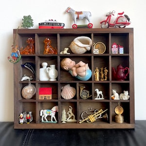 Puede incluir: Una caja de sombra de madera con 25 compartimentos llenos de varias figuritas y objetos en miniatura, incluyendo una muñeca bebé durmiendo, una tetera roja, una concha blanca, un caballo de balancín, un scooter rojo, una casita, un delfín, un par de jarras de cerámica blanca y una trompeta dorada.
