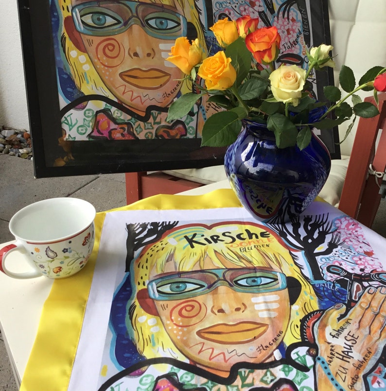 Pode incluir: Uma toalha de ch&aacute; amarela e branca com uma ilustra&ccedil;&atilde;o colorida do rosto de uma mulher com o texto "Kirsche Sonne Blumen" e "Nahezu fahren zu Hause". A toalha de ch&aacute; est&aacute; dobrada e colocada em uma mesa com um vaso azul de rosas amarelas.