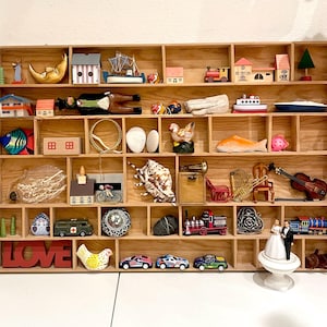 Könnte beinhalten: Ein Holzregal mit Miniatur-Dekorationsartikeln. Die Sammlung umfasst Häuser, Fahrzeuge, Musikinstrumente und Figuren. Das Regal ist aus hellem Holz gefertigt und an einer weißen Wand befestigt.