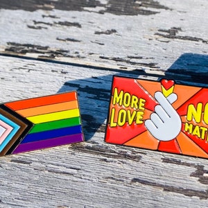 May include: Two enamel pins. The first pin is a rainbow flag with a black and brown triangle in the top left corner. The second pin is red with a white hand making a heart shape and the text "More Love No Hate".