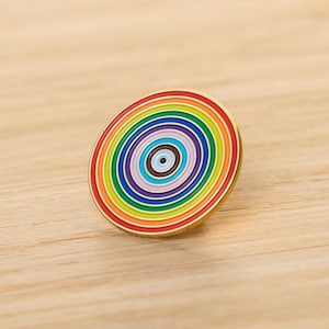 Puede incluir: Un pin redondo de esmalte con un diseño de círculos concéntricos de arcoíris. El pin tiene un borde dorado y está colocado sobre una superficie de madera. El diseño del pin presenta una serie de anillos coloridos, creando un accesorio vibrante.