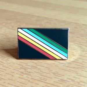 Könnte beinhalten: Eine rechteckige Emaille-Anstecknadel mit schwarzem Hintergrund und einem diagonalen Design aus farbigen Streifen. Die Streifen sind rot, gelb, weiß, türkis und grün. Die Nadel hat einen silberfarbenen Metallrand.