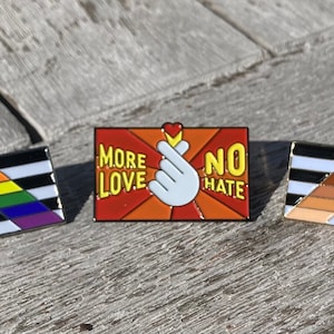 May include: Three enamel pins with different designs. The first pin is black and white with a rainbow triangle and the text "More Love No Hate". The second pin is red, orange, and yellow with a white hand making a heart shape and the text "More Love No Hate". The third pin is black and white with a brown triangle and the text "More Love No Hate".