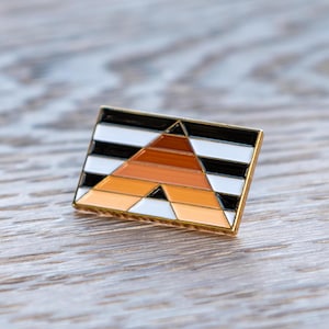 Puede incluir: Un pin de esmalte dorado con un diseño geométrico con rayas negras, blancas y naranjas. El diseño se asemeja a una pirámide con un triángulo negro en la parte superior.