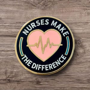 May include: A round metal pin with a black background and gold trim. The center features a light pink heart with a gold EKG heartbeat line. The text NURSES MAKE THE DIFFERENCE is printed in white capital letters around the heart, with two teal bars.