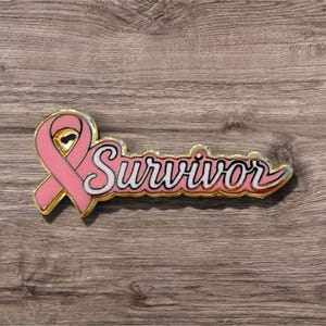 May include: A gold-toned metal enamel pin featuring a pink awareness ribbon on the left side, connected to the word Survivor written in a white cursive script with a pink outline and a gold-toned metal border, creating a cohesive design.