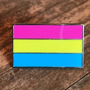 Könnte beinhalten: Eine silberne Anstecknadel mit einem horizontalen dreifarbigen Design. Das obere Band ist pink, das mittlere Band ist gelb und das untere Band ist blau.