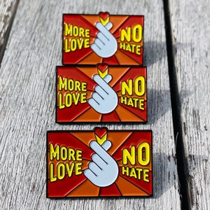 Puede incluir: Tres pines de esmalte con un esquema de color rojo, naranja y amarillo. Cada pin presenta una mano blanca haciendo un corazón con el texto "More Love No Hate" en amarillo.