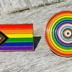 Może przedstawiać: Dwie szpilki emaliowane z wzorami tęczy. Pierwsza szpilka to prostokątna flaga z czarnym trójkątem, brązowym paskiem, jasnoniebieskim paskiem, różowym paskiem, białym paskiem i tradycyjnymi kolorami tęczy. Druga szpilka to okrągły wzór z koncentrycznymi okręgami w kolorach tęczy.