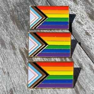 May include: Three enamel pins featuring a rainbow flag with a black and brown chevron in the corner. The pins are stacked on top of each other on a wooden surface.