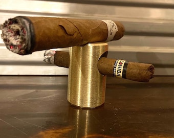 May include: A brass cigar holder with two cigars resting on it. The top cigar is lit and has a white label with black text. The bottom cigar has a black and red label with the text "COHIBA" and "REPUBLICA DOMINICANA".