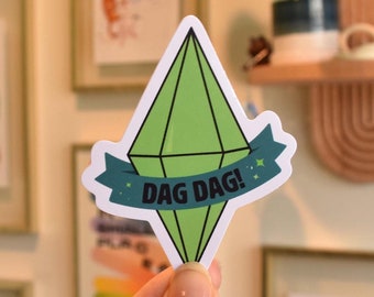 May include: A die-cut sticker shaped like a green diamond plumbob with a dark teal banner across the center featuring the text DAG DAG! in black capital letters. The sticker has a white border and small green star accents on the banner.