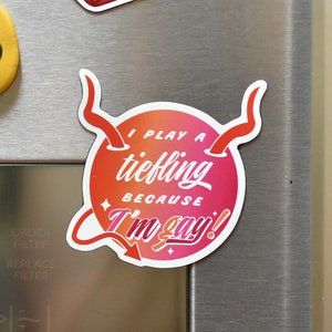 May include: A selection of magnets on a stainless steel surface. One is a yellow smiley face. Another is a red and orange die with the number 1. The third magnet has the text "I play a tiefling because I'm gay!"
