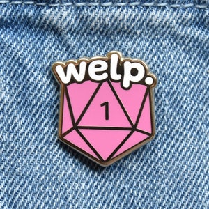 以下が含まれることがあります： 「welp.」という文字が書かれた、ピンクのエナメル製の二十面体のサイコロの形をしたピンバッジ。サイコロの面には「1」の数字が書かれている。