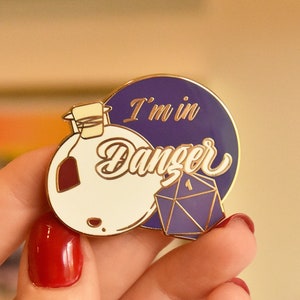 May include: A purple and gold enamel pin with a white potion bottle and a purple and gold dice. The pin says "I'm in Danger".